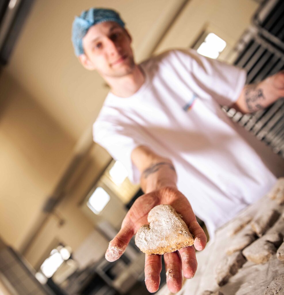 Produktions-Mitarbeiter hält ein Teigling eines Brötchens in Herzform zur Kamera hin in der Produktion.