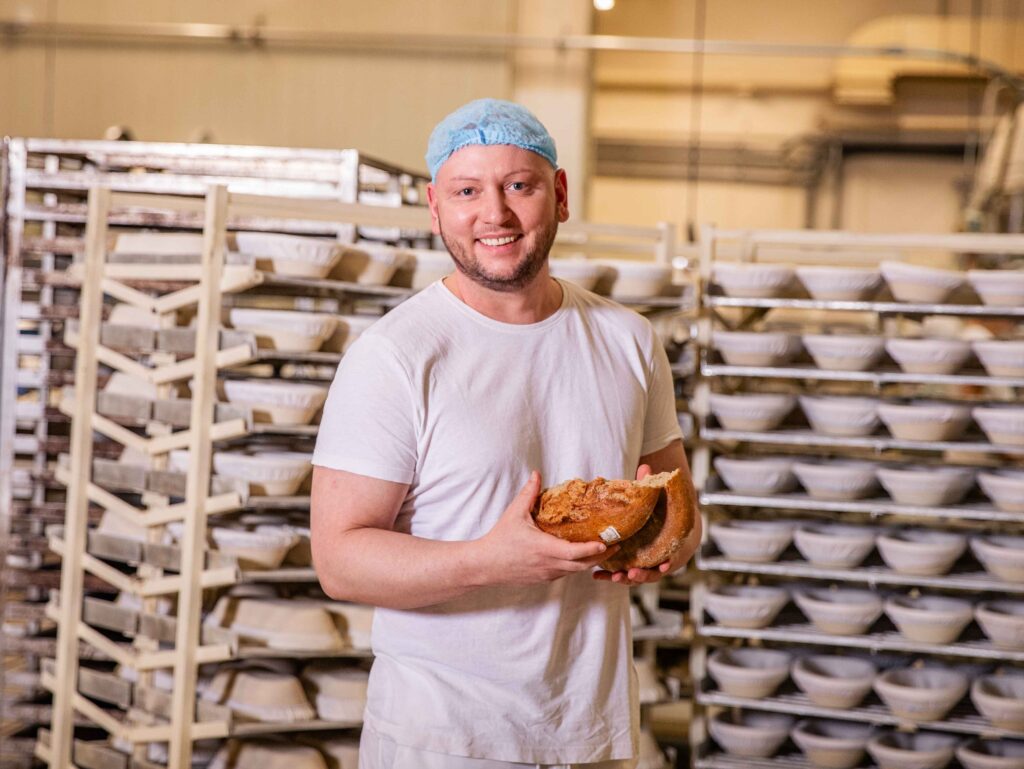 Bäcker in Arbeitskleidung bricht lächelnd ein Brotlaib in der Backstube. Im Hintergrund sind Stikkenwagen mit Brotkörben.