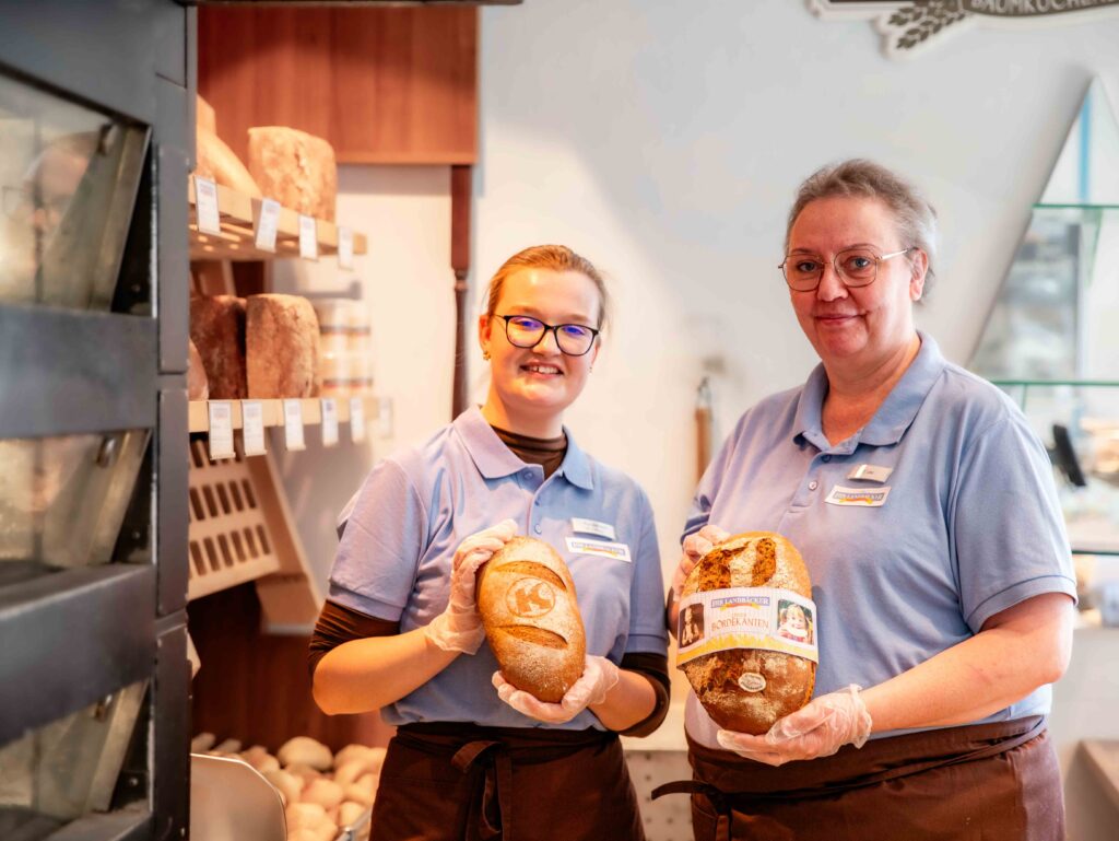 Zwei Bäckerei Fachverkäuferinnen, ca. 25 und 52 Jahre alt, stehen in Arbeitskleidung nebeneinander hinter der Theke einer Bäckerei Filiale und halten bei jeweils ein Brotlaib in den Händen. Sie schauen Lächelnd in die Kamera.