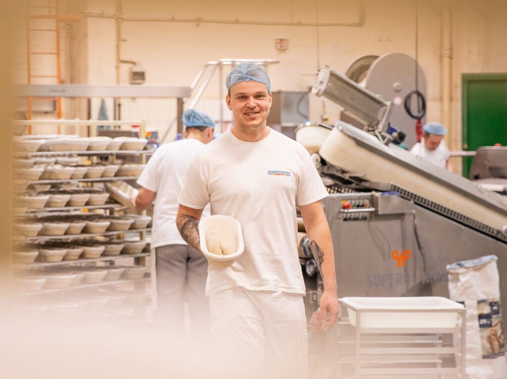 Bäcker trägt in Arbeitskleidung einen Brotkorb mit einem Brot-Teigrohling durch die Produktion.