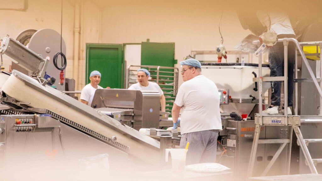 Drei Bäcker, im Alter von 29-52 Jahren, arbeiten in Arbeitskleidung, an einer großen Maschine in der Produktion.