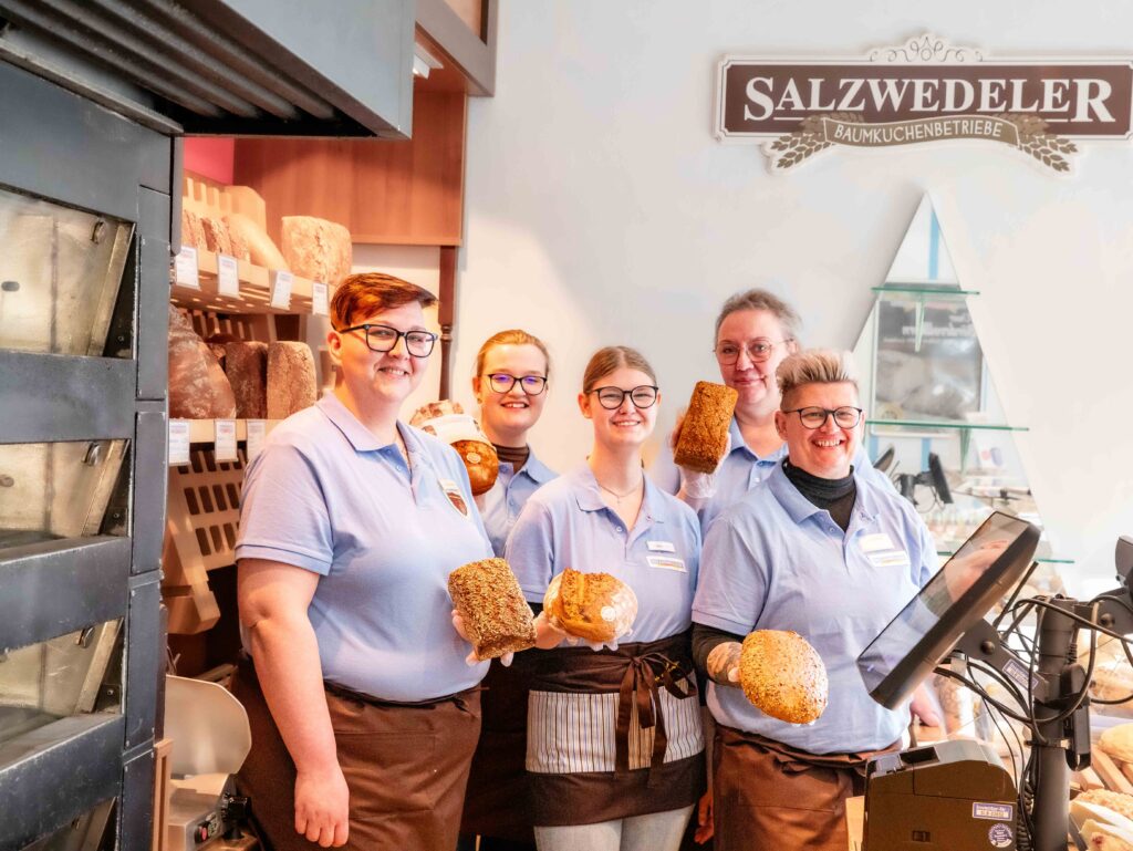 Fünf Bäckerei Fachverkäuferinnen stehen in einer Gruppe zusammen in einer Bäckerei Filiale und tragen dabei Arbeitskleidung. Alle lächeln in die Kamera und halten jeweils unterschiedliche Brote in der Hand.