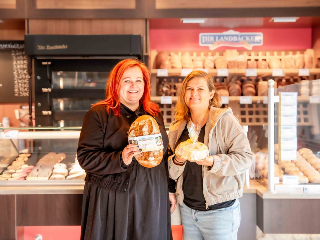 Die beiden Geschäftsführerinnen vom Stendaler Landbäcker, Anja Bosse (ca.35 Jahre alt) und Kristin Dessert (ca. 32 Jahr alt), stehen nebeneinander in einer Bäckerei Filiale. Anja hält ein Brotlaib und Kristin ein Salzwedel Baumkuchen. Beide lächeln in die Kamera.