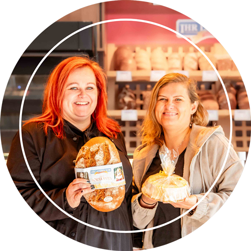 Die beiden Geschäftsführerinnen vom Stendaler Landbäcker, Anja Bosse (ca.35 Jahre alt) und Kristin Dessert (ca. 32 Jahr alt), stehen nebeneinander in einer Bäckerei Filiale. Anja hält ein Brotlaib und Kristin ein Salzwedel Baumkuchen. Beide lächeln in die Kamera.