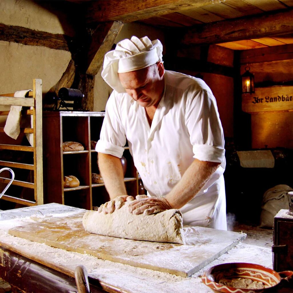 Bäcker und Gründer von Ihr Landbäcker Andreas Boss, in Traditioneller Bäckerkleidung in einer rustikalen Backstube am Teig kneten.