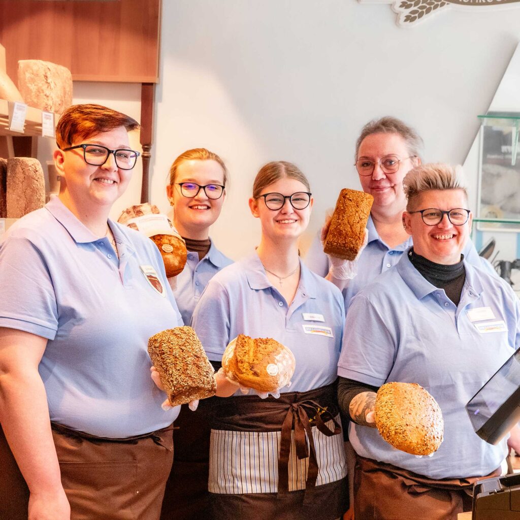 Drei Fachverkäuferinnen, eine Praktikantin und eine Auszubildende stehen in einer Ihr Landbäcker Filiale und posieren lächelnd mit einem Brotlaib in der Hand.