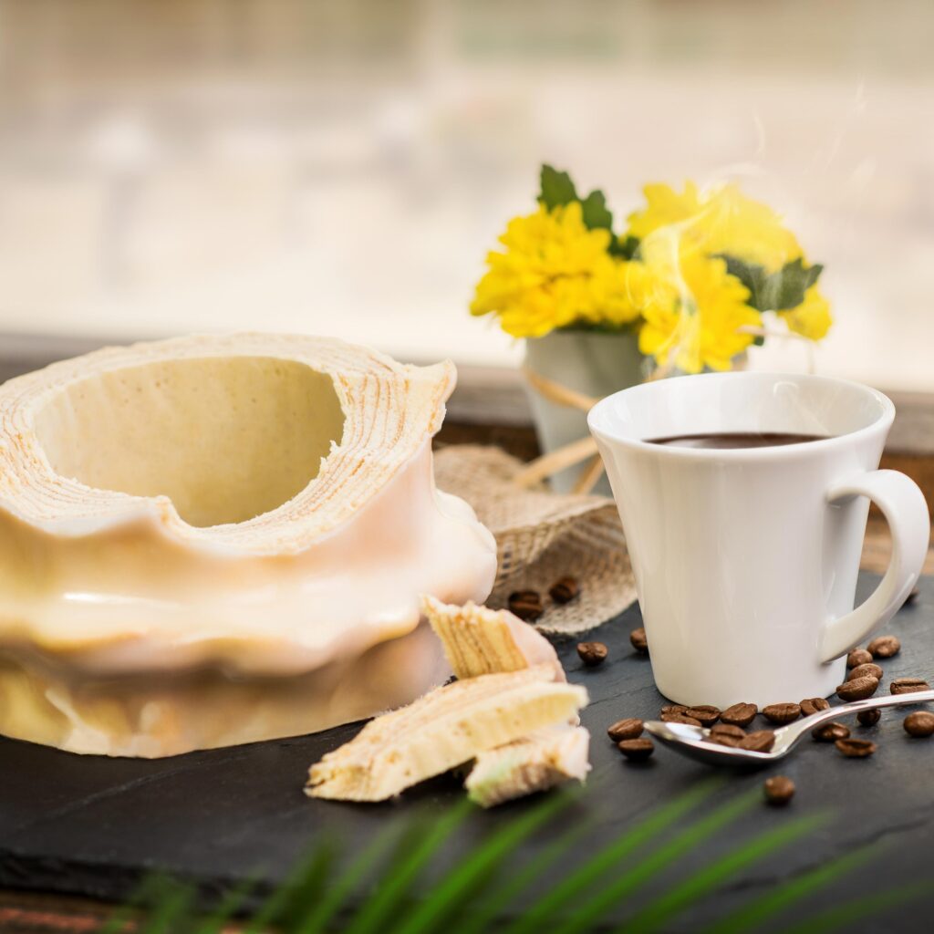 Weißer Baumkuchen aufgeschnitten schön drapiert mit einer kleinen Kaffeetasse und Blumen im Hintergrund.